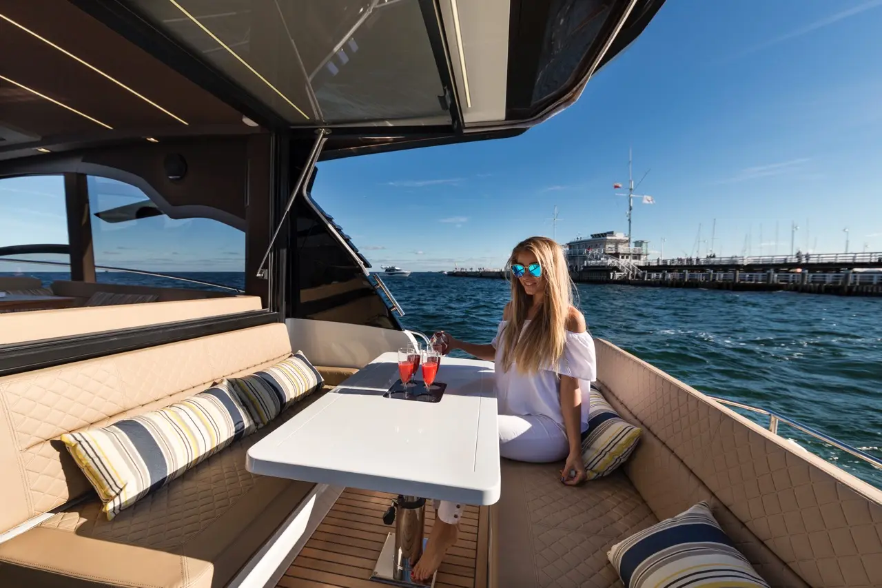 Woman in sunglasses enjoying cocktails on a luxury boat with plush seating and a view of a marina in the background.