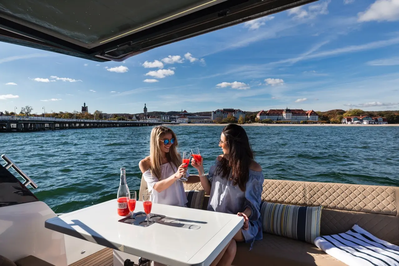 Two women toast with drinks on a boat, with a scenic waterfront and buildings in the background on a sunny day.