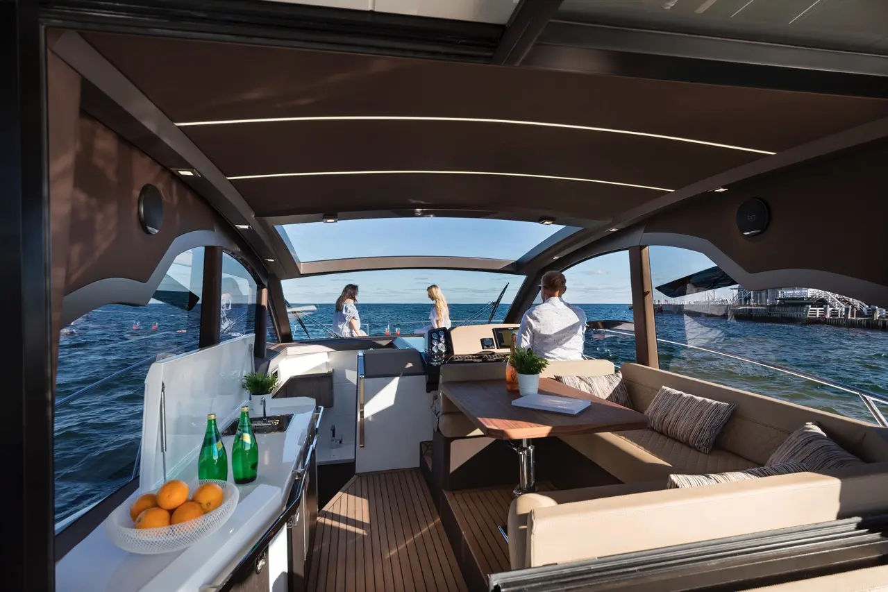 Interior view of a luxury boat with a table, drinks, and three people enjoying the ocean view through large windows.