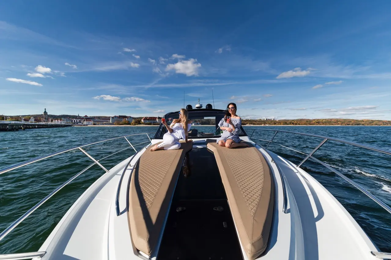 Two women relax on a luxury boat's sunbeds, enjoying drinks with a scenic waterfront view and clear blue skies.