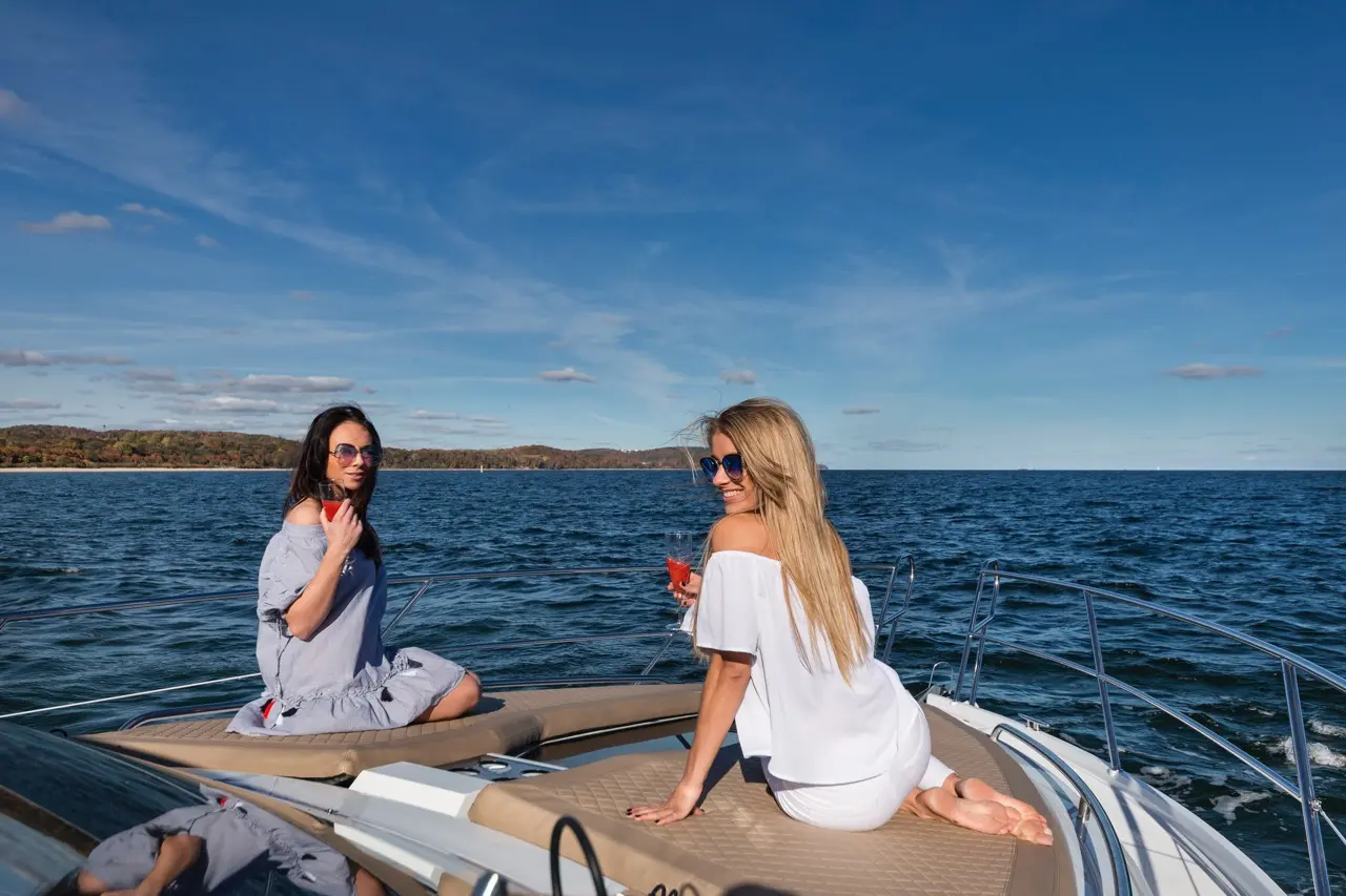 Two women relax on a boat, enjoying drinks. One wears a white off-shoulder top, the other a blue dress, against a scenic backdrop.