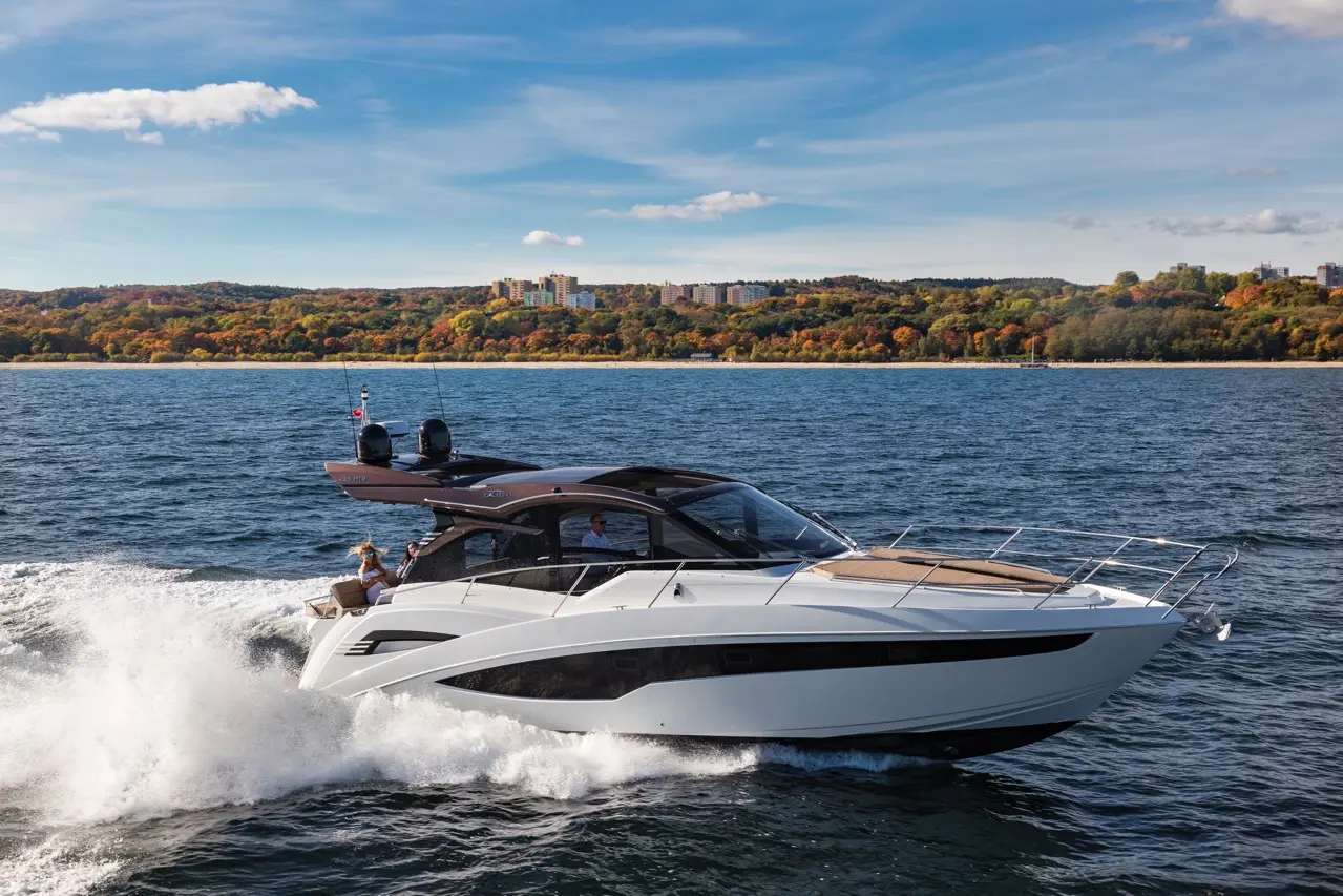 A luxury boat speeds across the water, with a backdrop of colorful autumn trees and buildings under a clear blue sky.