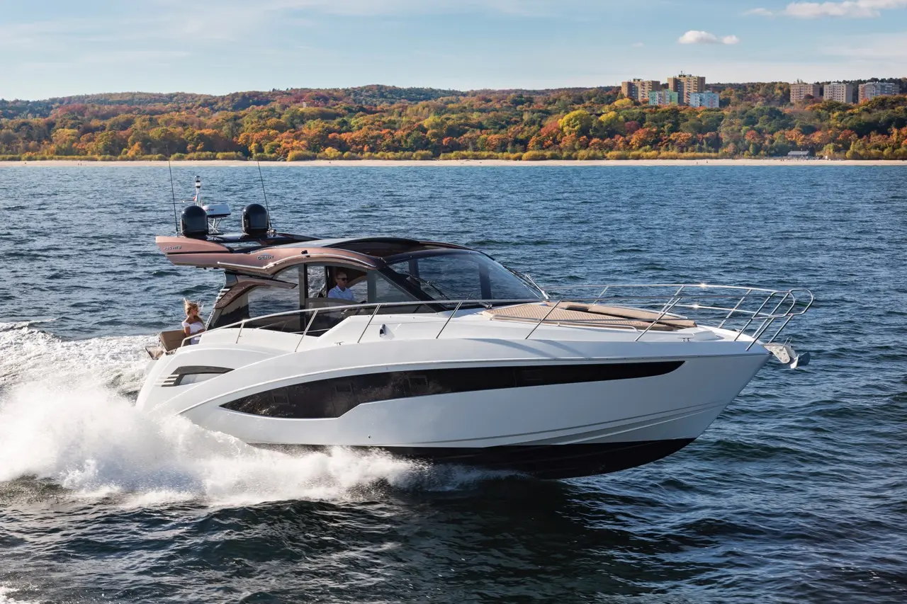 A sleek white motorboat speeds across the water, surrounded by autumn foliage and a distant shoreline under a clear sky.