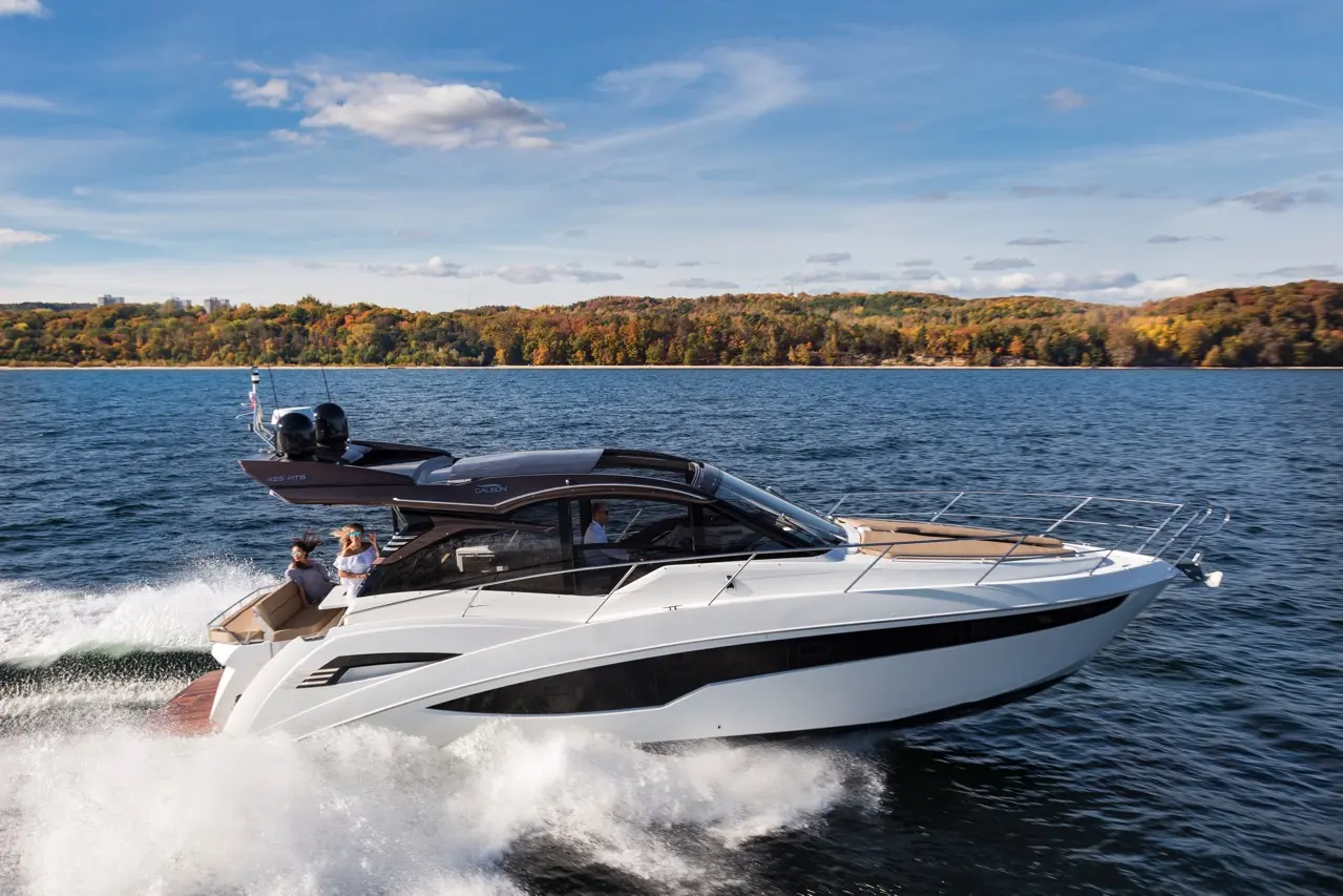 A sleek white motorboat speeds across a lake, with two people enjoying the ride while surrounded by autumn foliage.