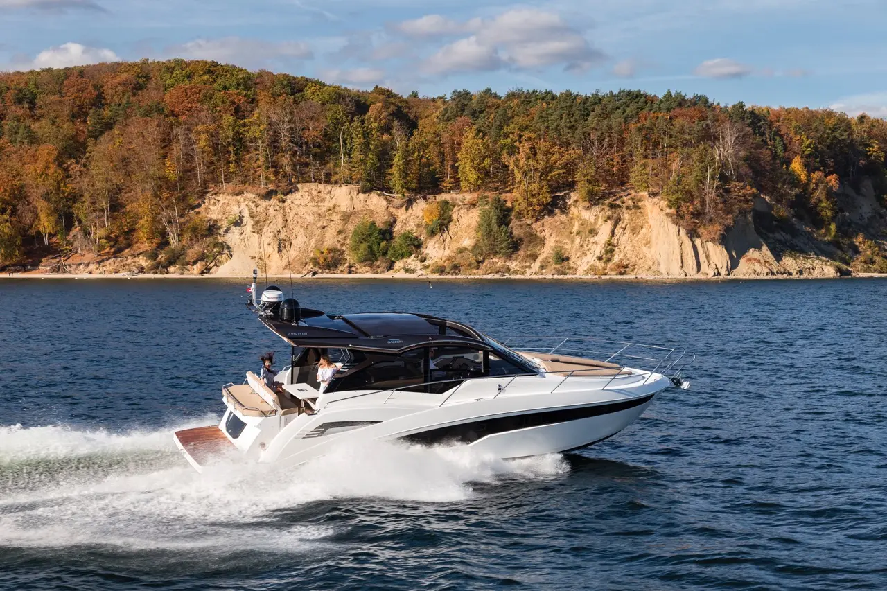 A sleek white motorboat speeds across sparkling water, with a scenic autumnal forest backdrop and a sandy shore.