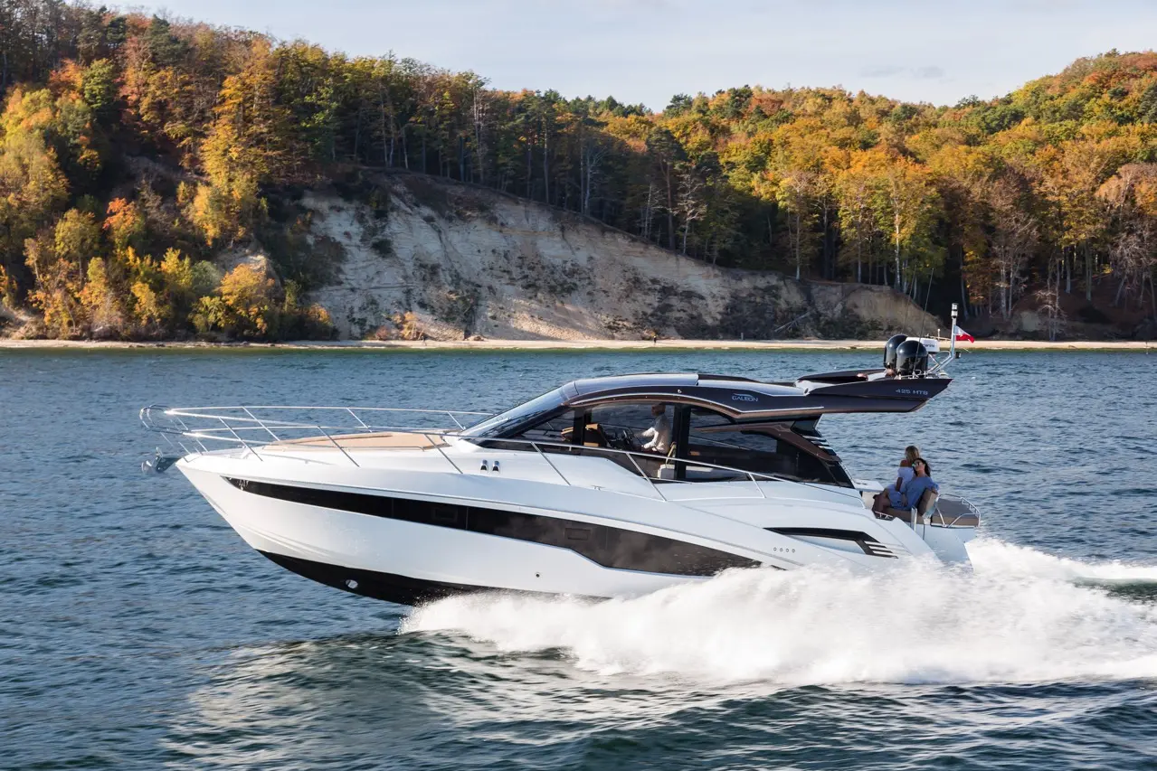 A sleek white motorboat speeds across clear water, surrounded by colourful autumn trees and a sandy shoreline.