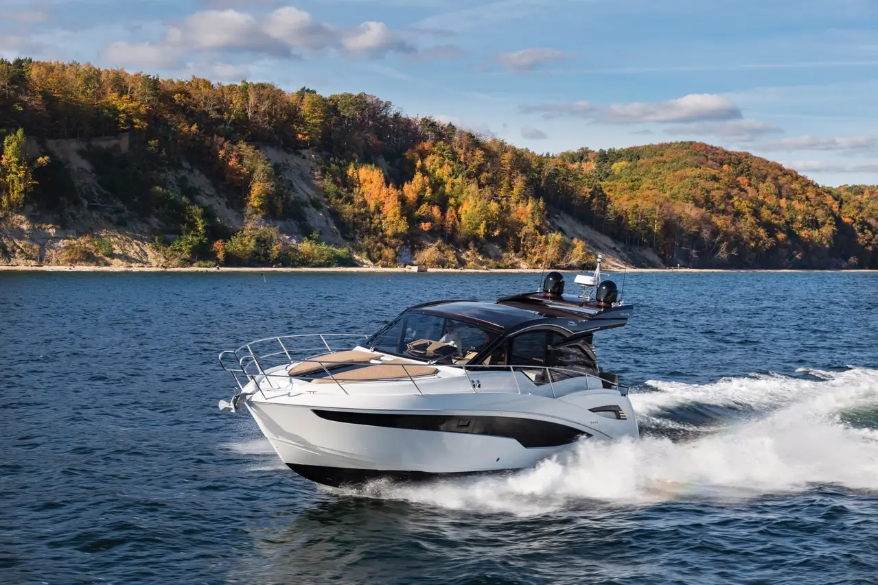 A white motorboat speeds across a blue lake, surrounded by autumnal trees on a rocky shore under a clear sky.