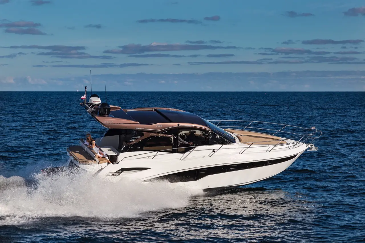 A sleek white motor yacht speeds across the blue sea, creating splashes, with a person enjoying the ride on board.