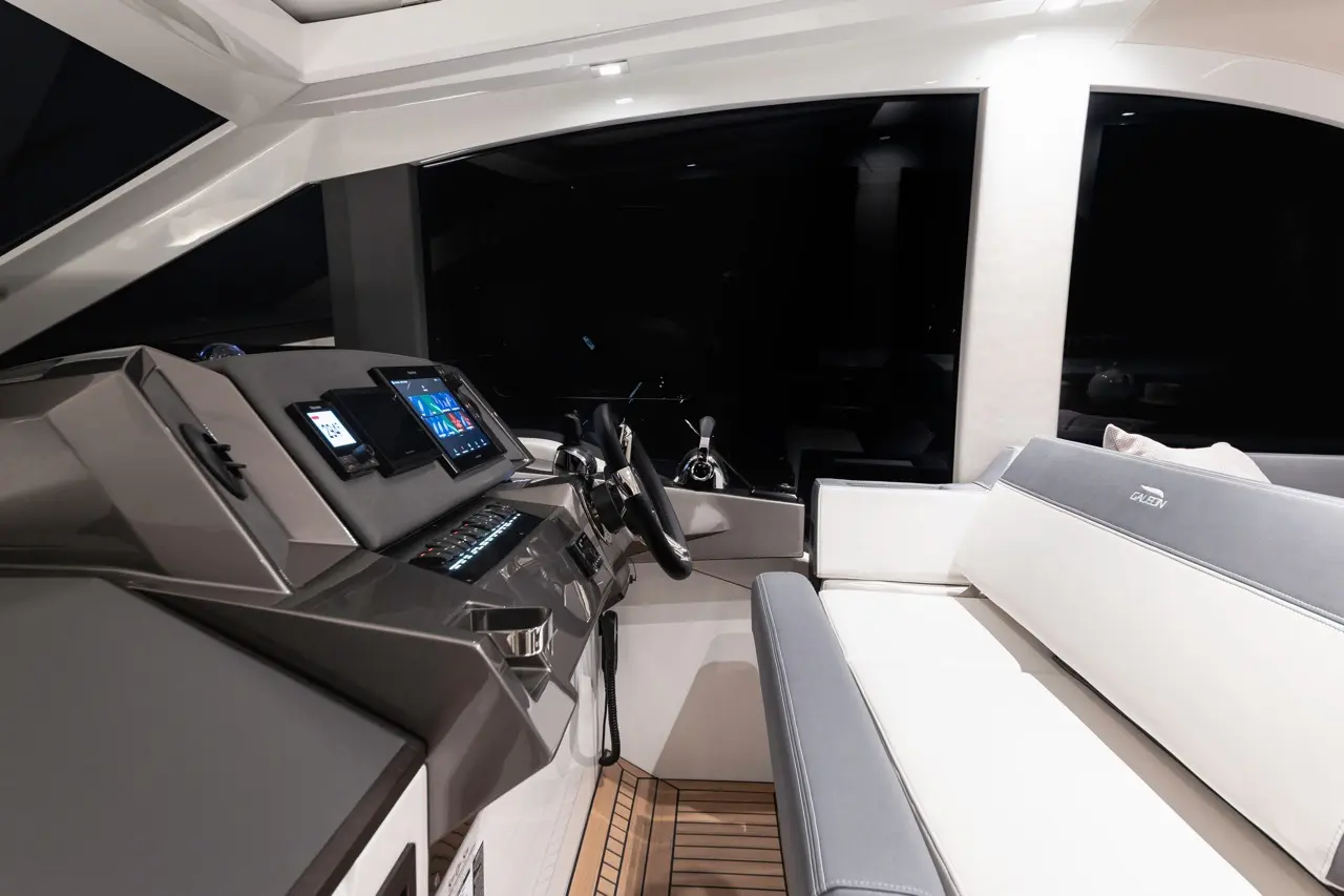 Luxury yacht interior featuring a modern helm with screens, steering wheel, and white seating on wooden flooring.