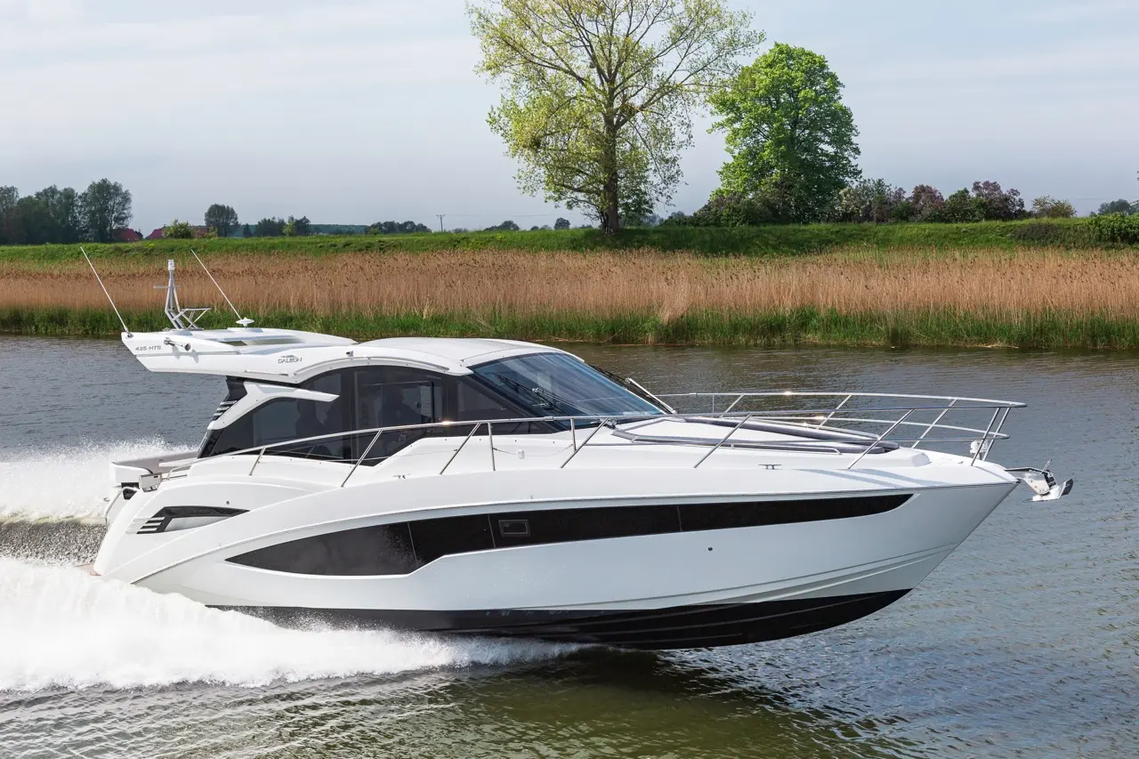 A sleek white motorboat glides through calm waters, with green trees and tall grasses lining the shore.