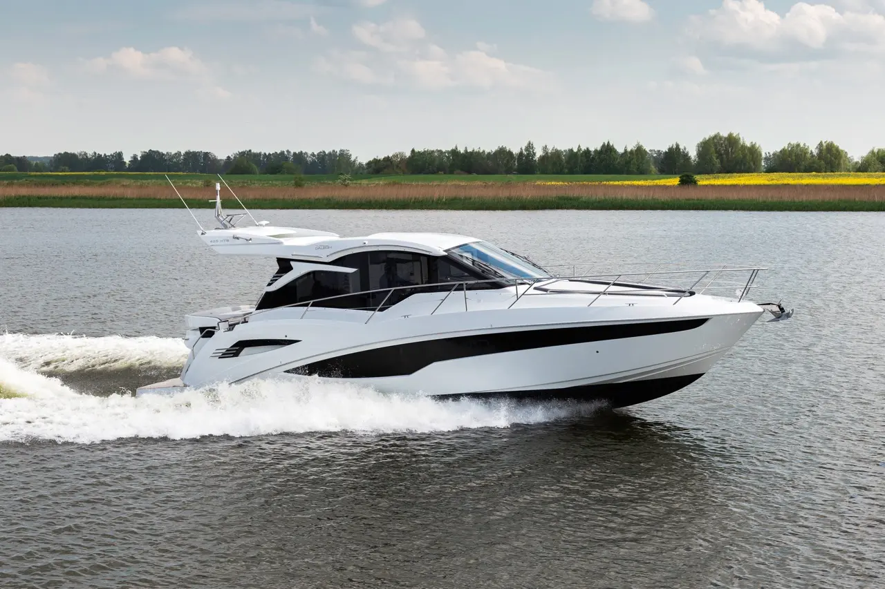 A sleek white motorboat glides across calm water, with green hills and a field of yellow flowers in the background.