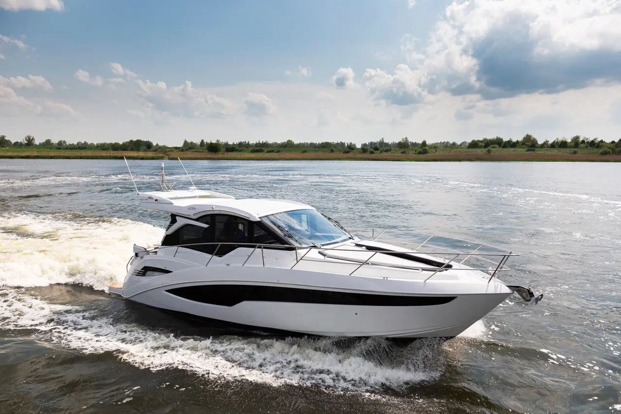 White luxury motorboat cruising on a river, generating waves under a blue sky with fluffy clouds and lush greenery in the background.