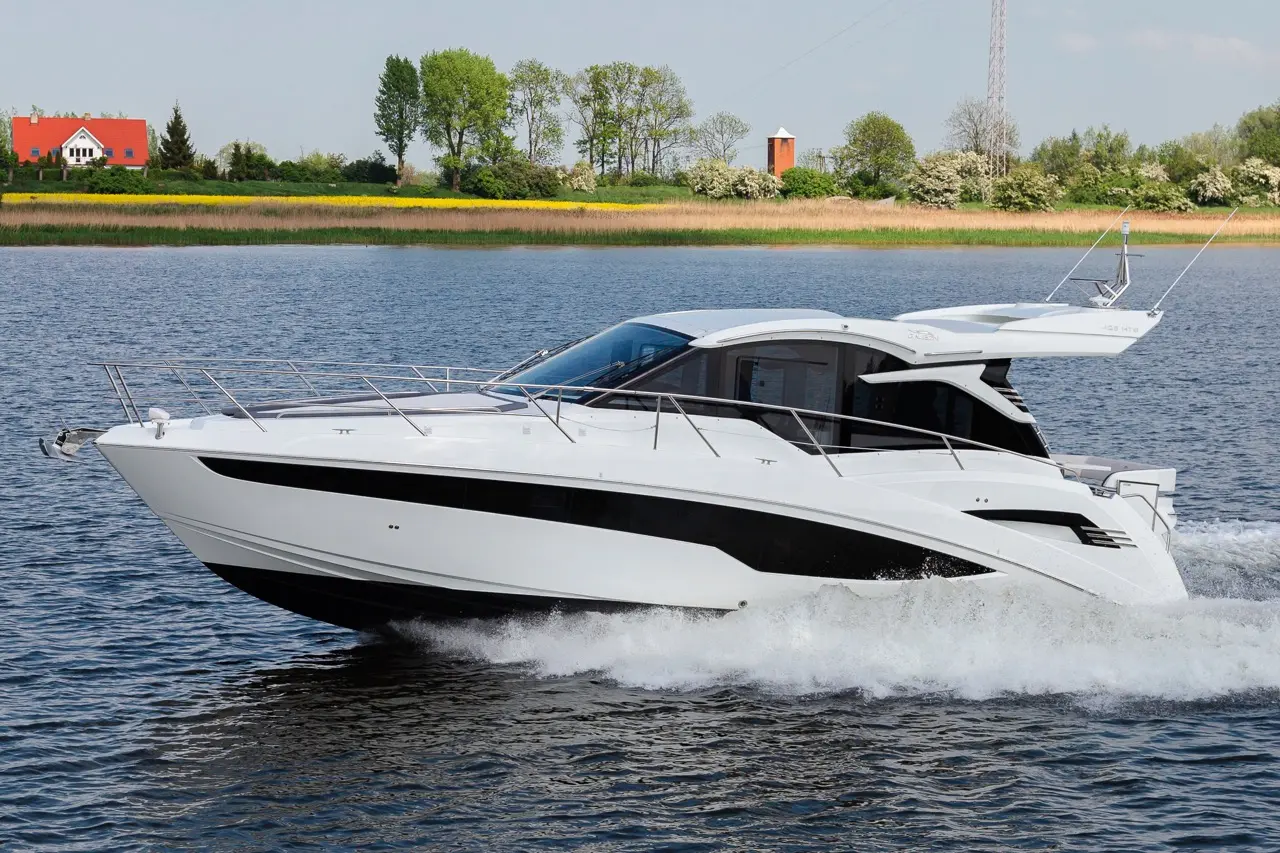 A sleek white motor yacht cruising across calm water, with green trees and a distant house in the background.