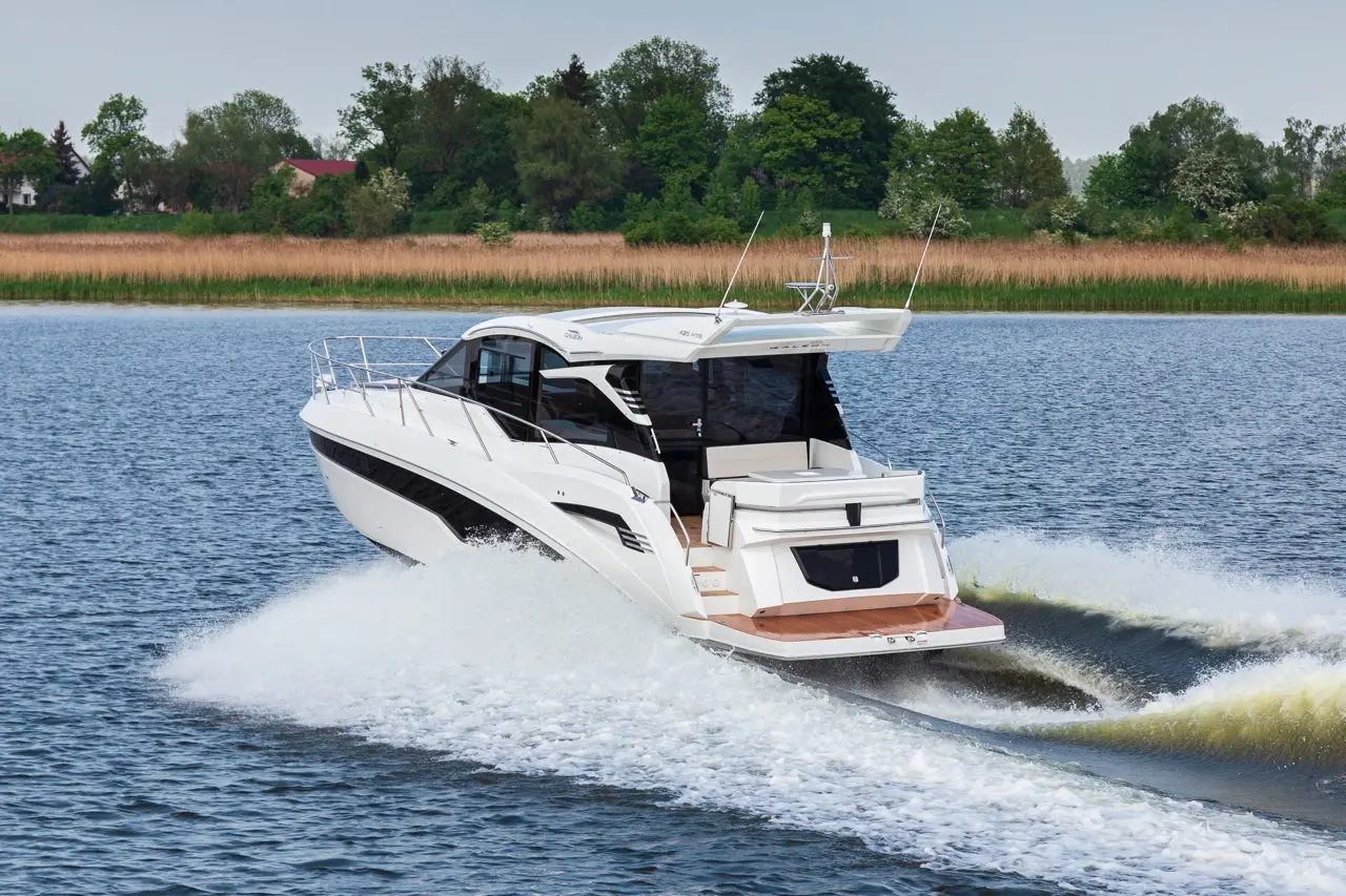 A luxury motorboat speeds across a lake, leaving a wake of white foam against a backdrop of lush greenery and reeds.