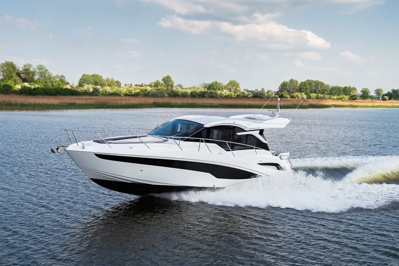 A sleek white yacht glides over calm water, leaving a frothy wake, with green trees lining the shore under a blue sky.