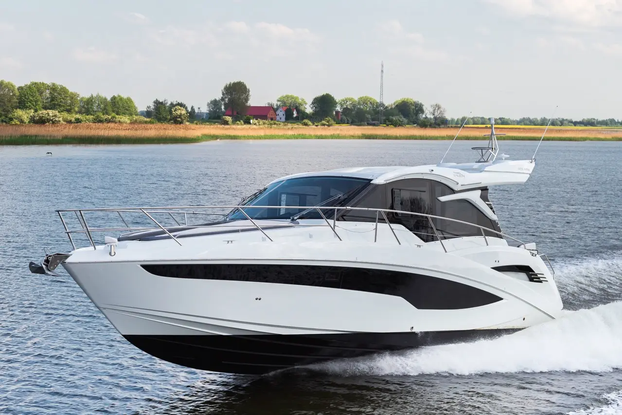 A sleek white motorboat speeds across calm water, with lush green vegetation and distant houses in the background.