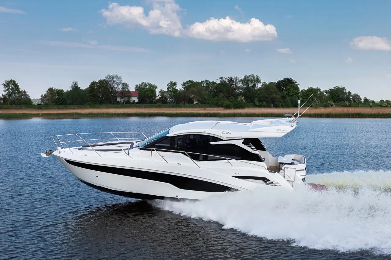 Luxury motorboat cruising on calm waters, leaving a wake, with a lush green shoreline and blue sky in the background.