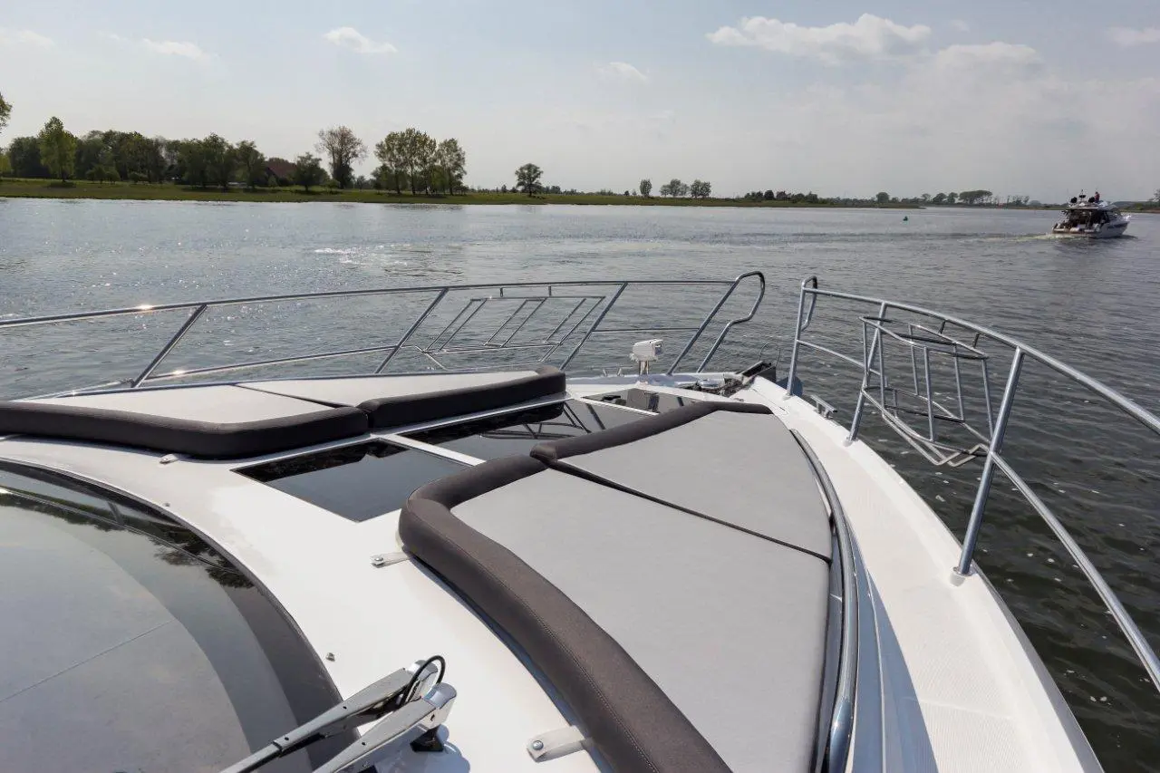 View from the bow of a luxury boat with grey cushions, overlooking a calm river and greenery under a sunny sky.