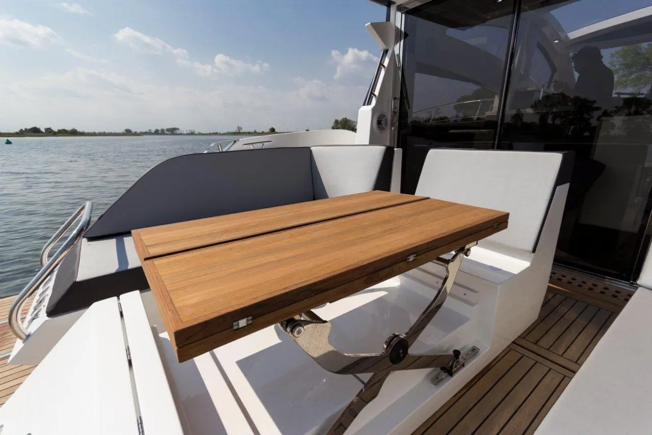 Sunlit yacht deck features a wooden table with folding mechanism, surrounded by white seating and a serene water backdrop.
