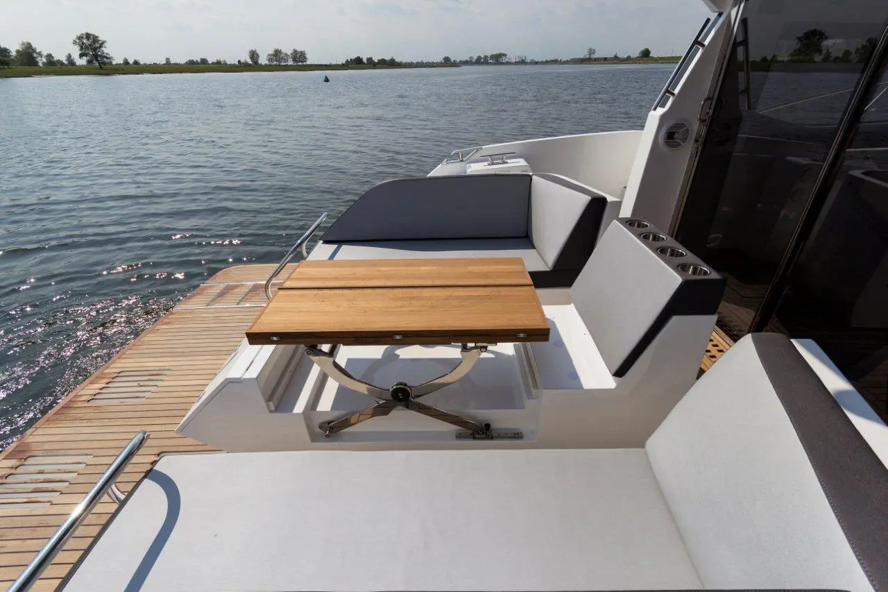 A modern boat deck featuring a wooden table, comfortable seating, and views of the calm water with trees in the background.