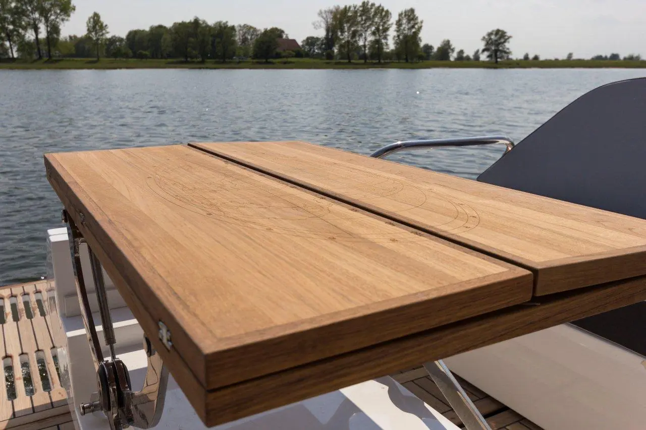 Teak table mounted on a boat deck, overlooking a lake with trees in the background and a clear sky above.