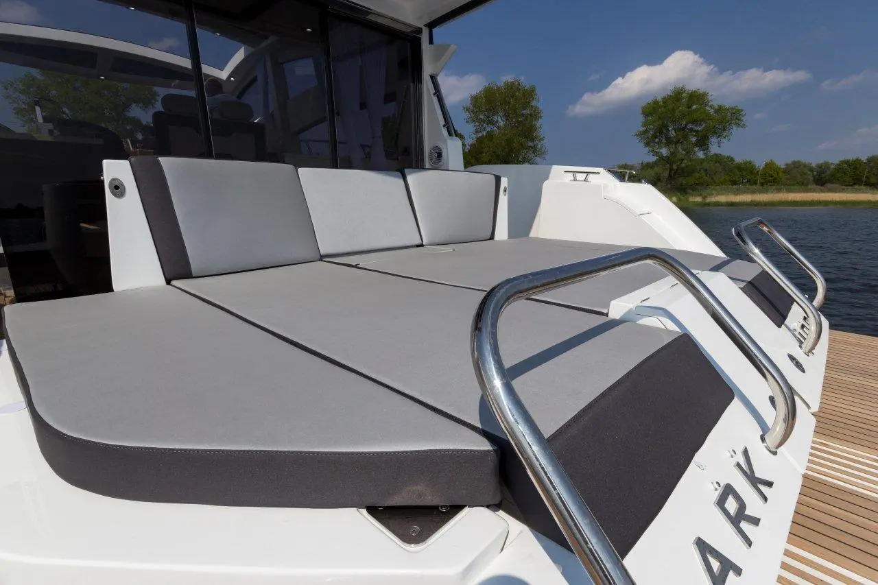 Lounge area on a modern boat with gray cushions, stainless steel rails, and views of water and trees under a blue sky.