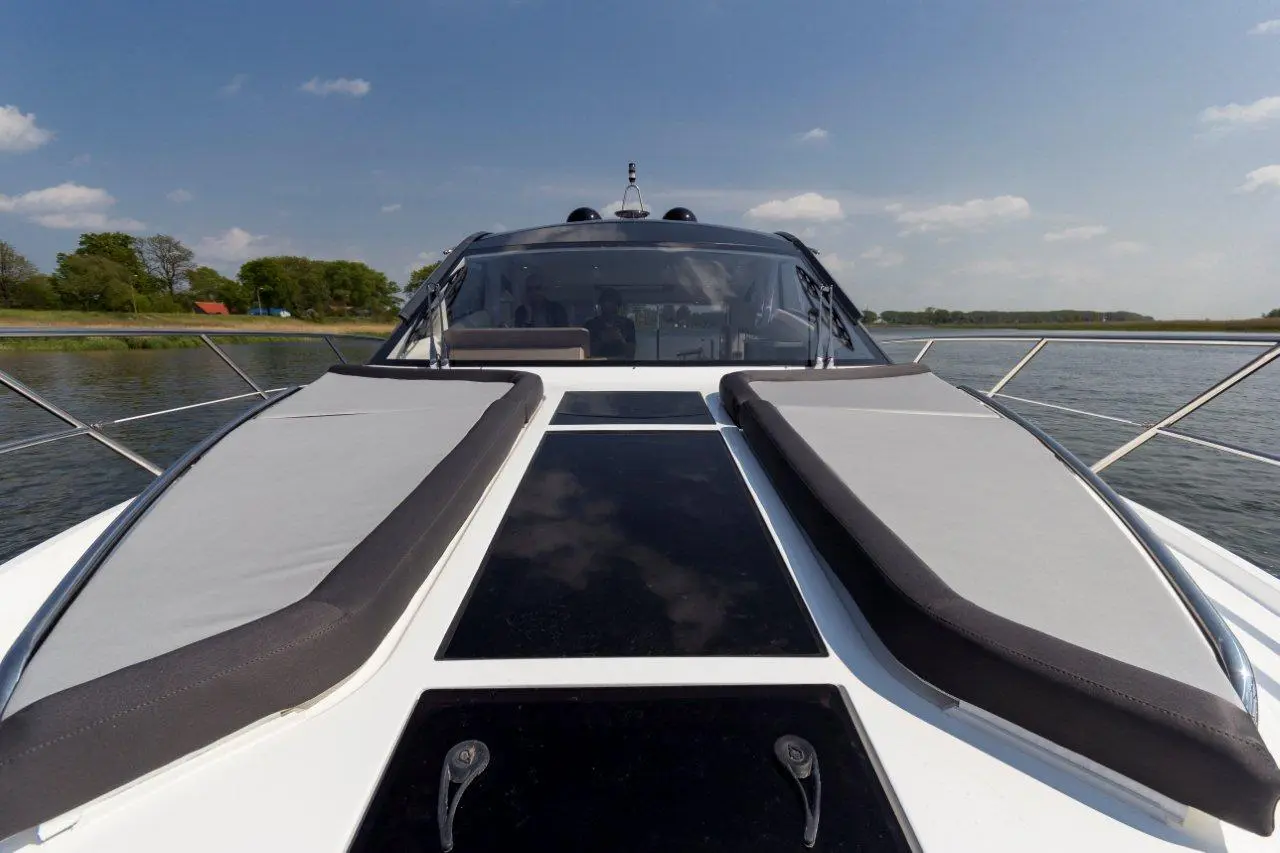 A view from the front of a white motorboat showing cushioned sunbeds and a scenic riverside landscape under a blue sky.
