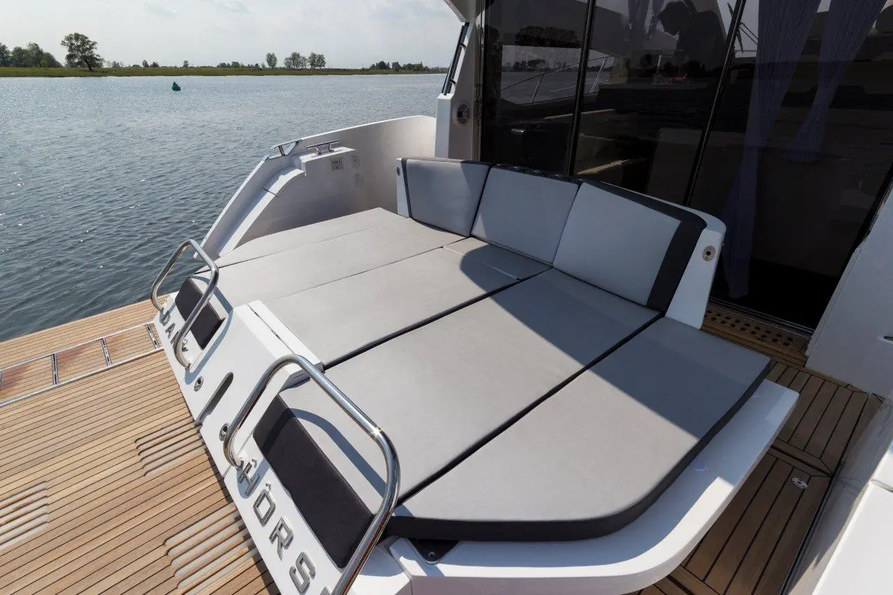 A yacht's stern features gray cushioned seating, overlooking a calm waterway with lush green trees in the background.