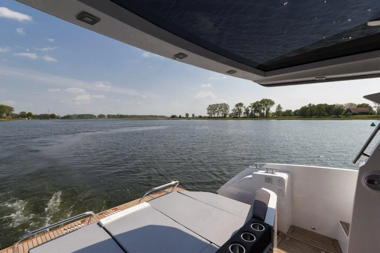 View from a boat featuring cushioned seating, a metal railing, and calm water reflecting a clear sky and green trees.