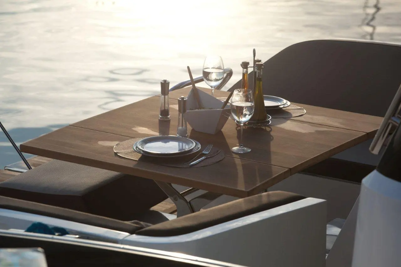 Elegant outdoor dining setup on a boat, featuring wooden table, glassware, cutlery, and olive oil. Sunlight reflects on water.