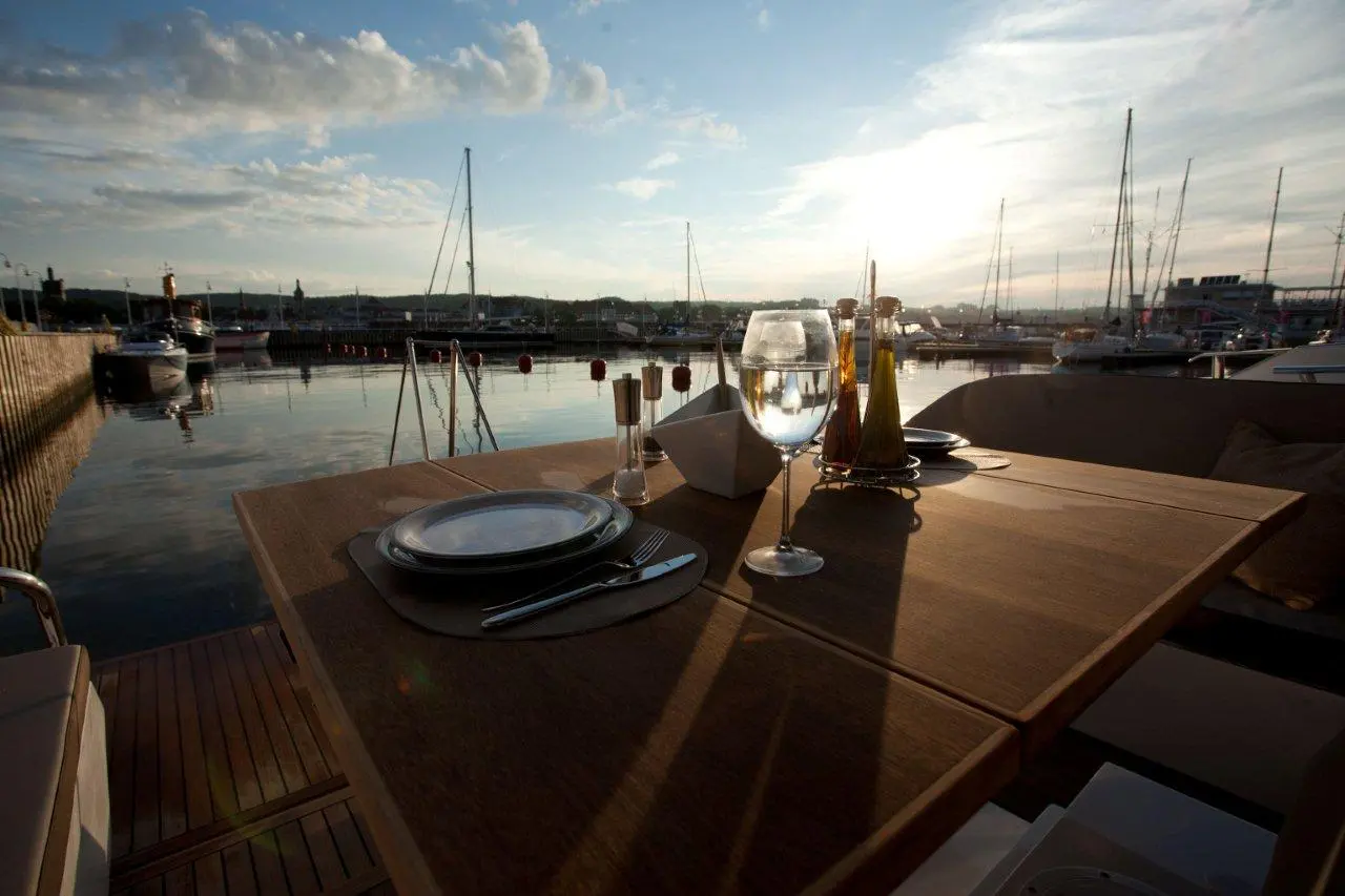 A scenic marina at sunset with a wooden table set for dining, featuring glassware, condiments, and reflections on the water.