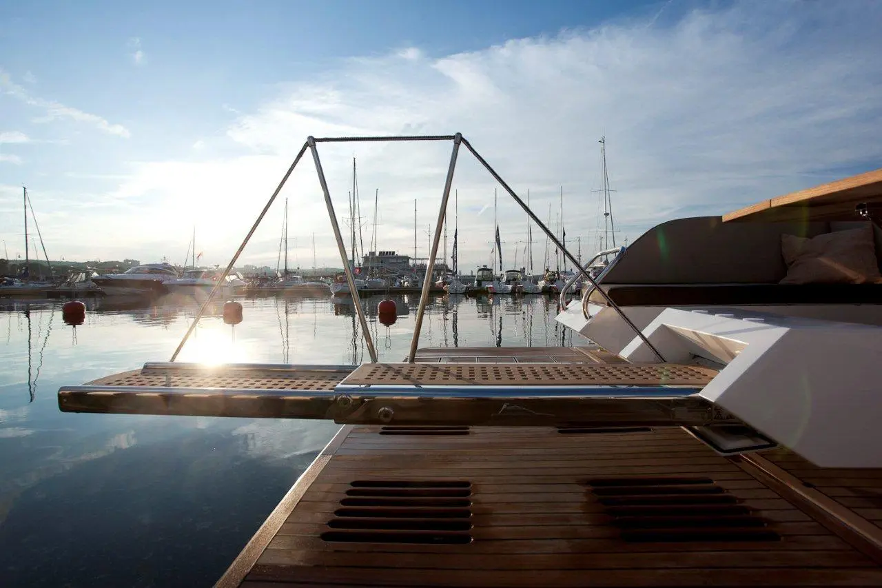 A sleek boat platform extends over the calm water, reflecting a marina filled with sailboats under a bright sky.