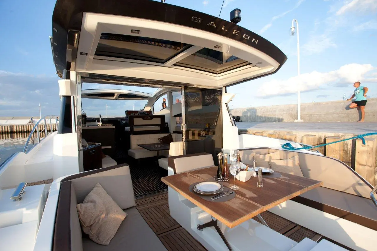 Luxury yacht interior with seating, dining setup, and a person stretching on the dock in the background under a blue sky.
