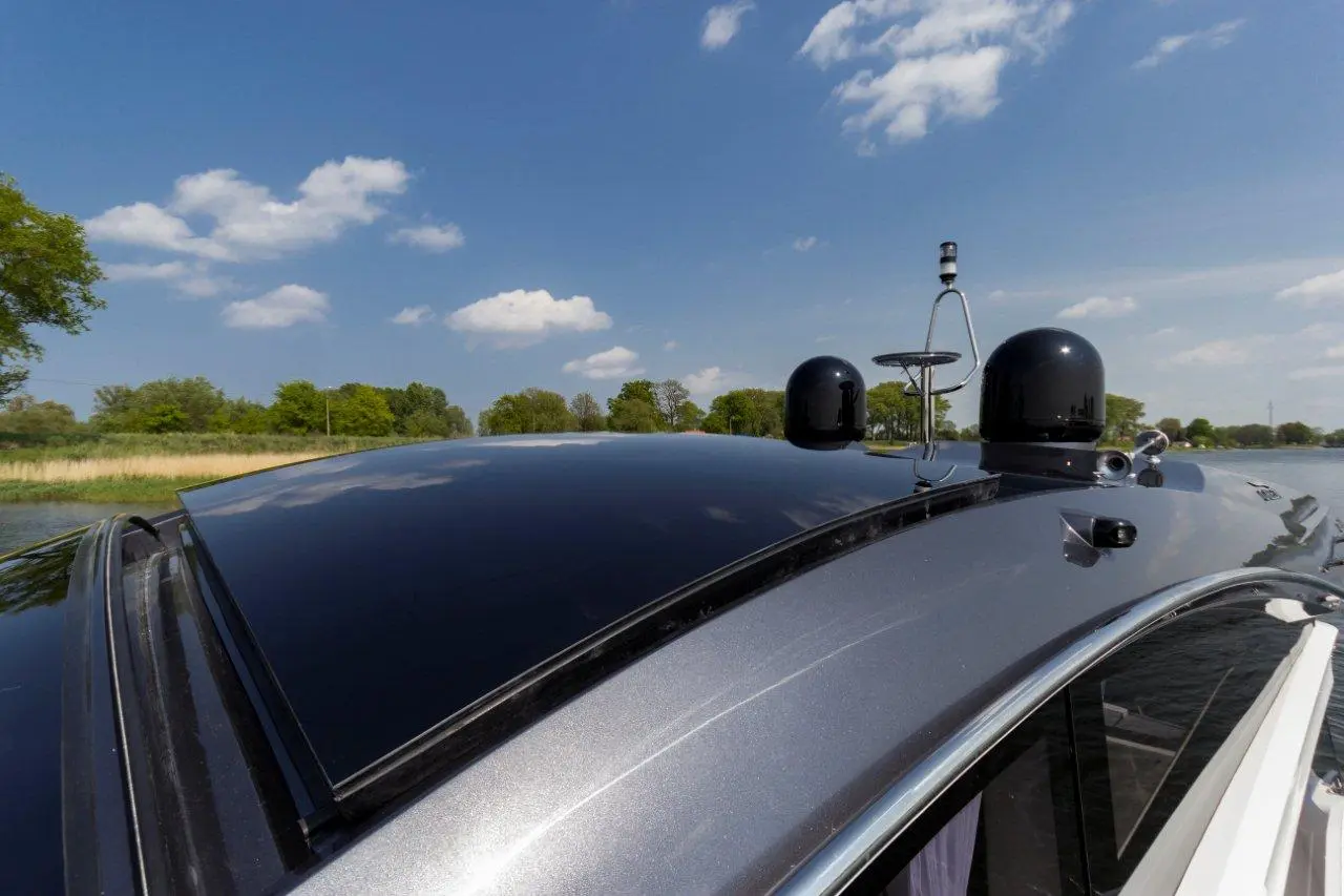 A close-up view of a boat's shiny silver roof, showing antennas against a blue sky and green trees by a waterway.