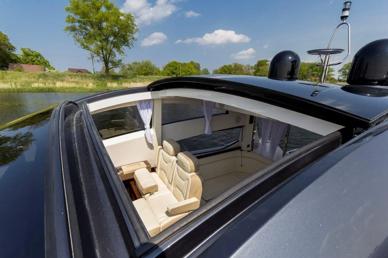 Luxury boat interior with beige seating and windows, overlooking a calm river and greenery under a blue sky.