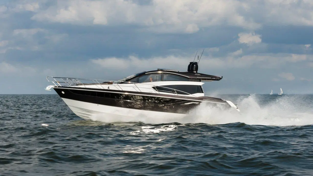 A sleek motor yacht speeds across a choppy sea under a cloudy sky, with a small sailboat visible in the distance.