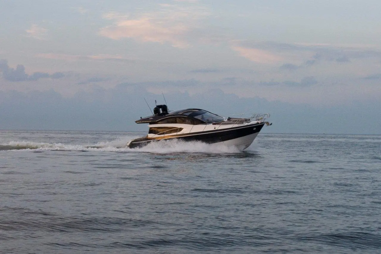 A sleek motorboat speeds across calm waters under a cloudy sky, creating ripples in its wake.