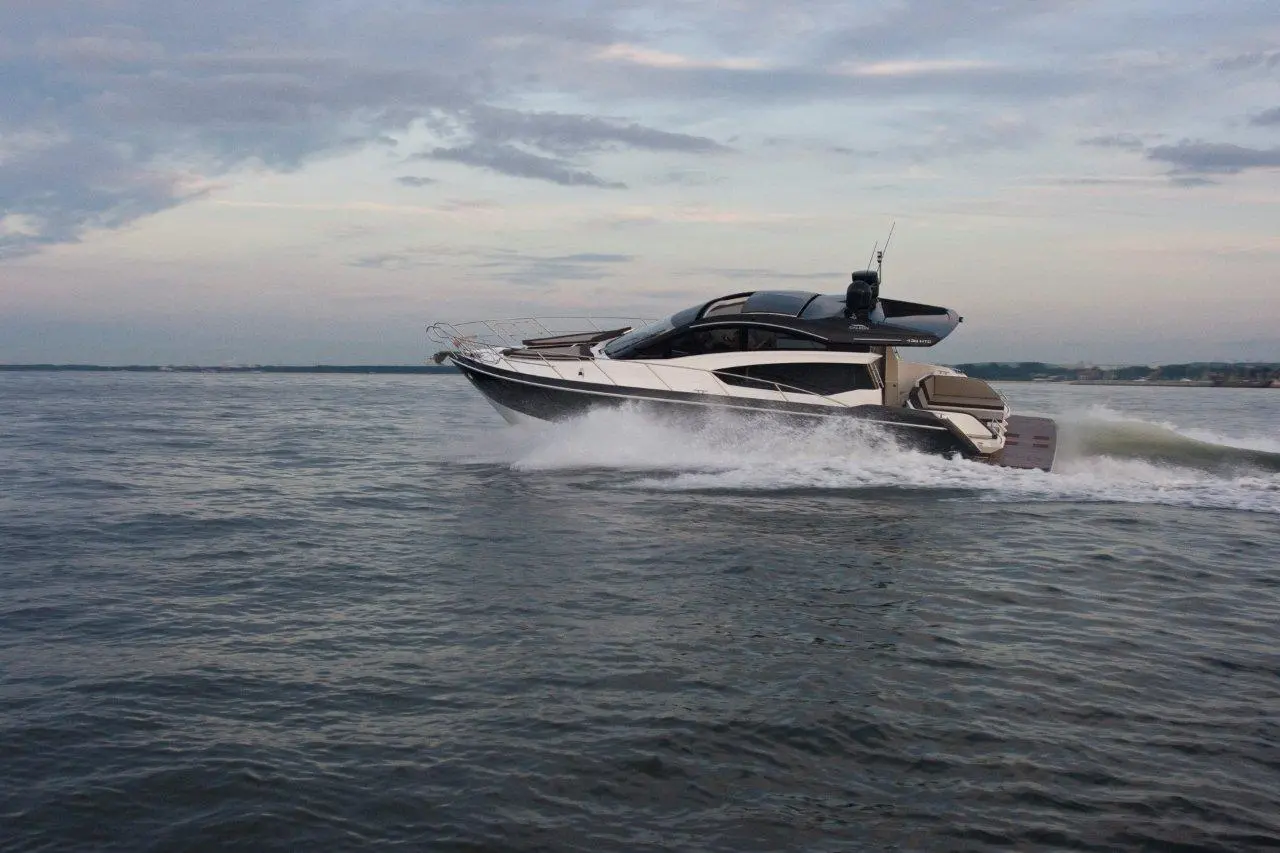 A sleek motor yacht glides across calm waters, creating splashes as it speeds along under a cloudy sky.