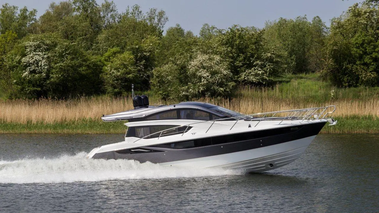 A sleek white and black motorboat speeds across a calm lake, surrounded by lush green trees and tall grass.