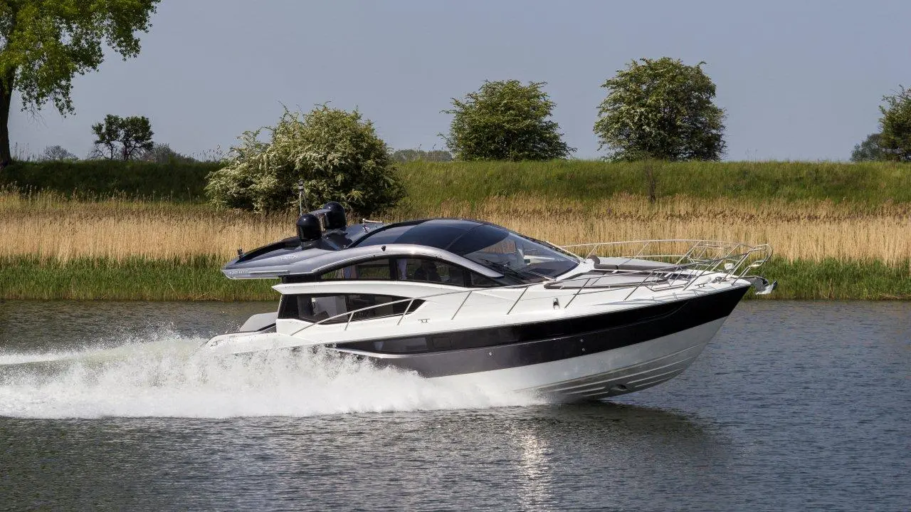 A sleek motorboat speeds through calm water, leaving a white wake behind, with green trees and grass in the background.