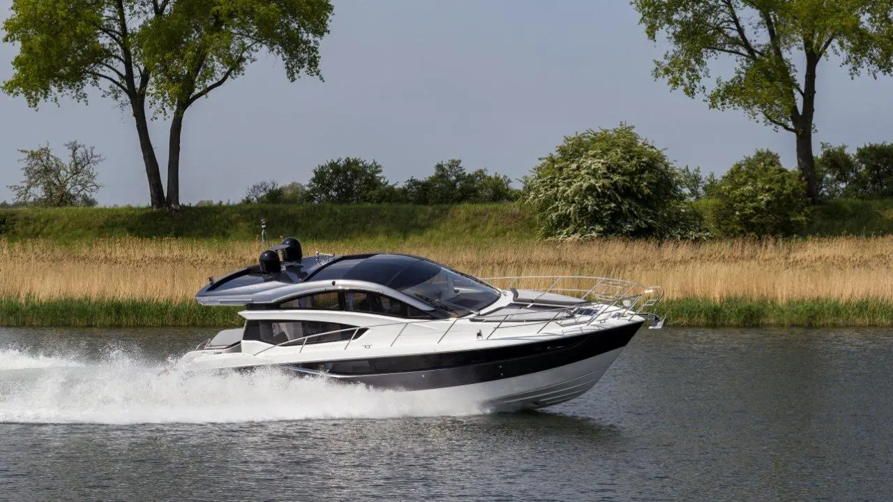 A sleek motorboat speeding across a calm river, surrounded by green trees and tall grass under blue skies.
