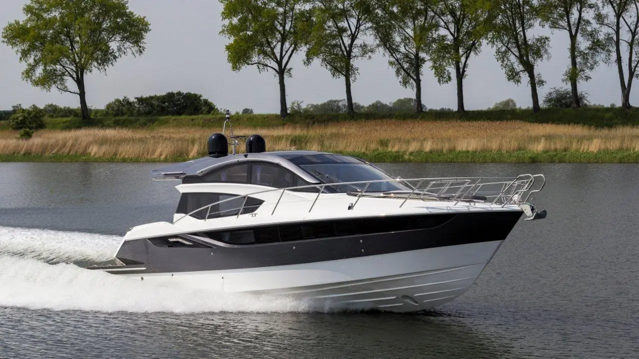 A sleek motor yacht glides through calm waters, framed by tall trees and grass, showcasing luxury and outdoor leisure.