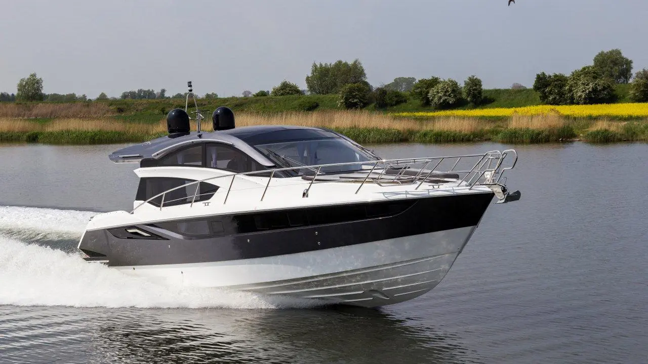 A sleek motorboat glides through calm waters, with greenery and yellow flowers lining the shore in the background.