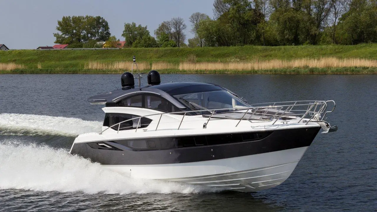 A sleek motorboat gliding through calm waters, with a green bank and trees in the background under a clear blue sky.