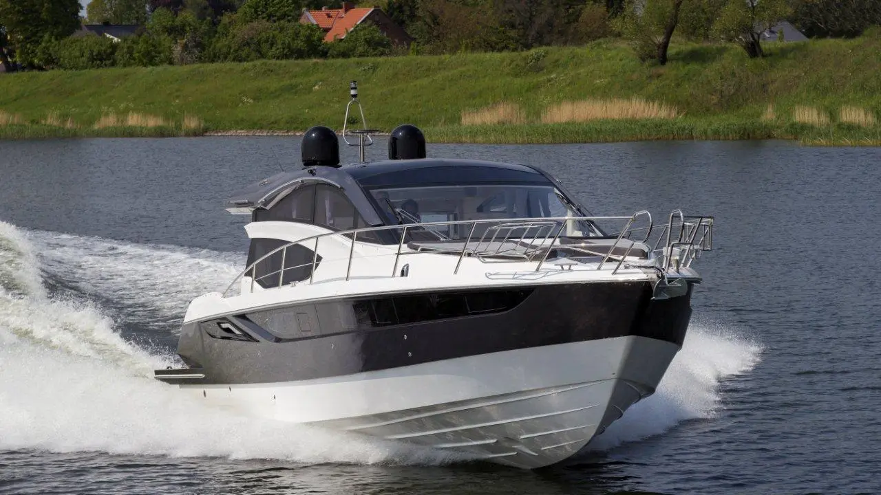 A sleek motor yacht speeds across a calm lake, creating a white wake against lush green banks and a clear blue sky.