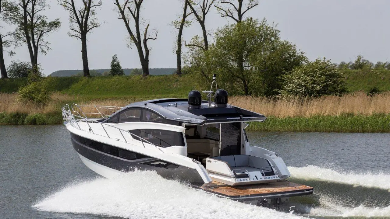 A sleek motorboat cruises on calm waters, creating a graceful wake against a backdrop of trees and greenery.