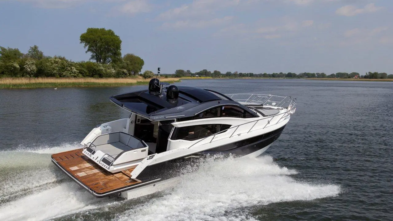 A sleek black and white motor yacht speeds across a calm river, creating a wake in the water with green trees in the background.