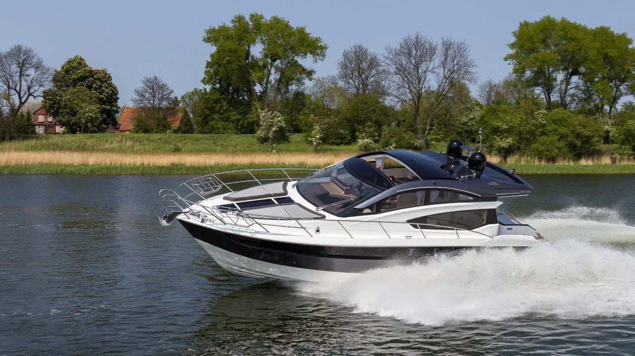 A sleek white and black boat speeds across a calm lake, with trees and a house visible in the background.