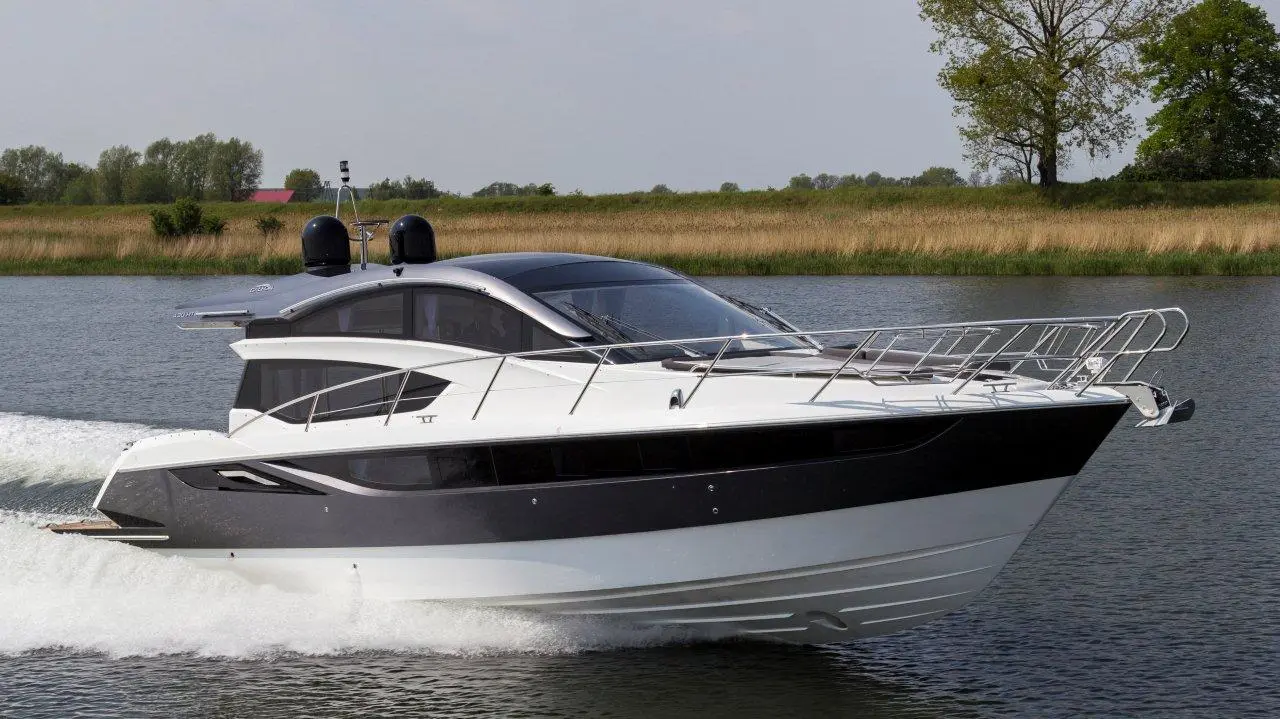 A sleek motor yacht glides through calm water, showcasing a modern design with black and white accents, surrounded by greenery.