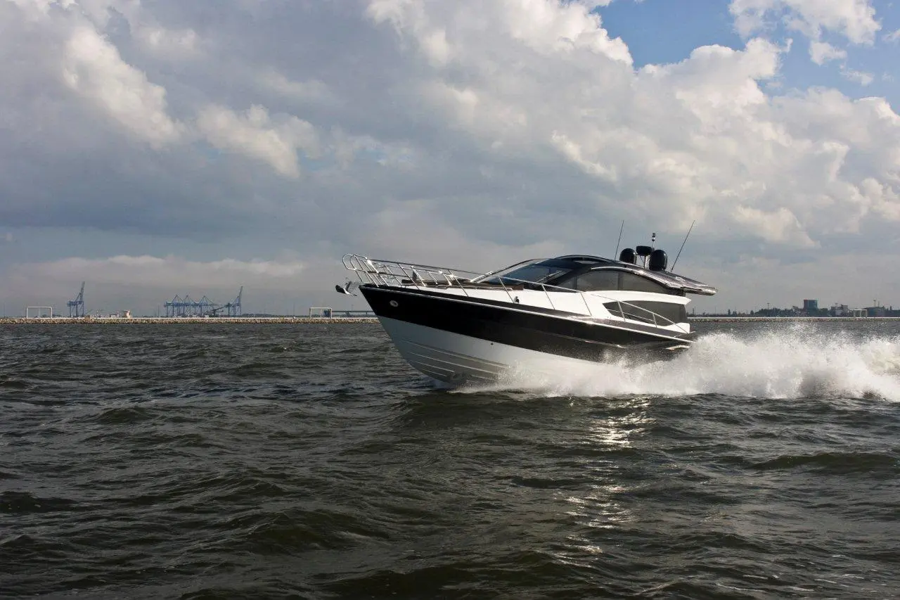 A sleek black and white speedboat navigates choppy waters under a cloudy sky, with a distant industrial backdrop.